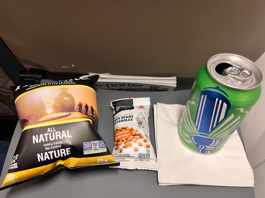 Porter Airlines onboard catering consisting of potato chips, baked fava beans, and a beer, on the seat-back tray table.